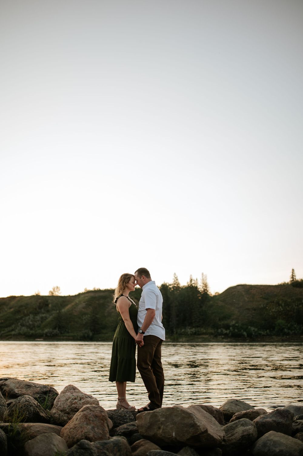 A couple stands on rocky shoreline against sunset backdrop with river and trees in background.