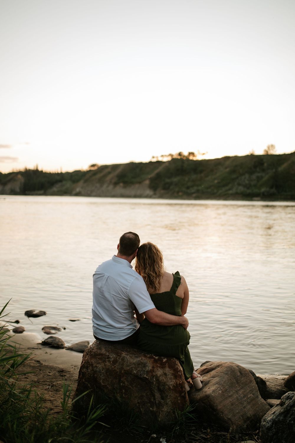 A couple sits together on a rocky shoreline at sunset overlooking a serene lake or river.