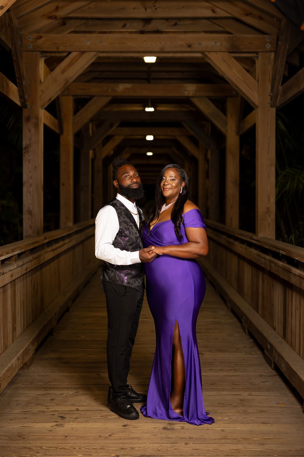A couple poses elegantly in a covered wooden bridge walkway, wearing formal attire.