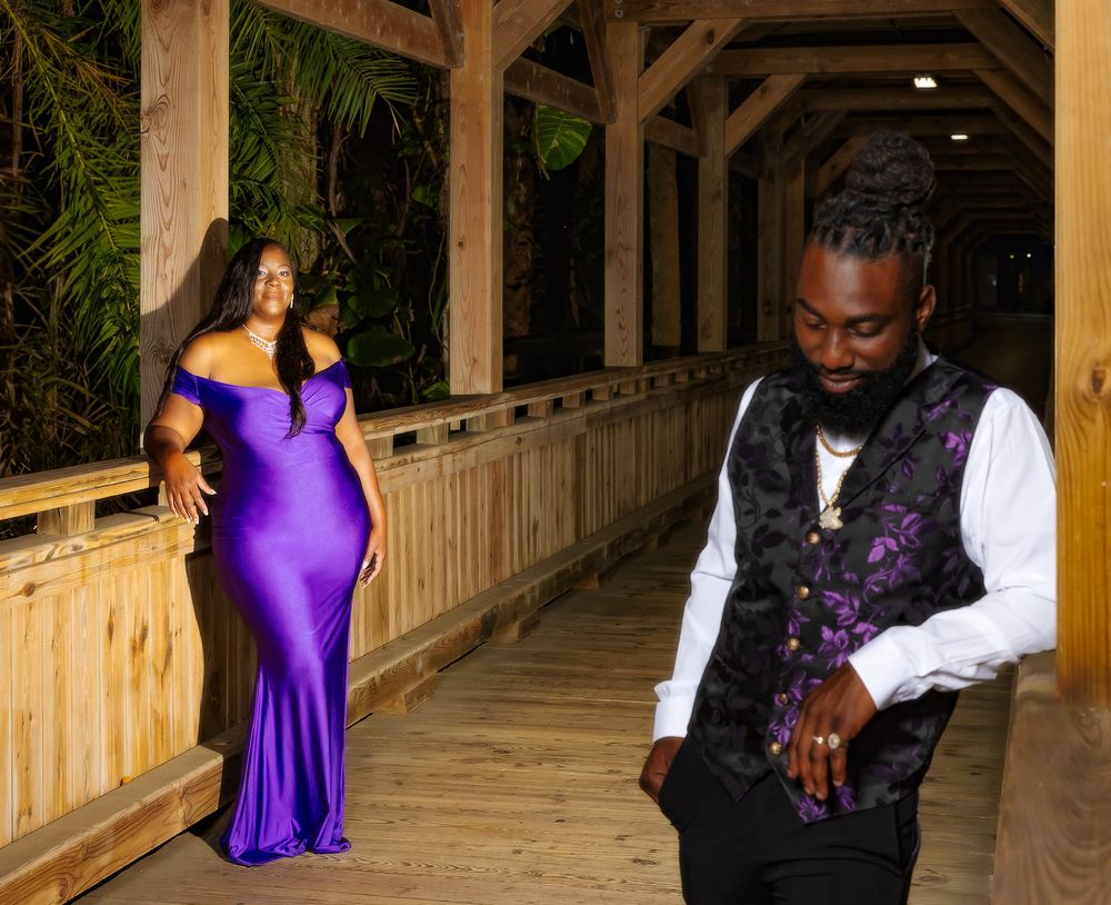 A couple poses on a wooden walkway with palm trees, wearing formal attire including a purple gown and dark vest.