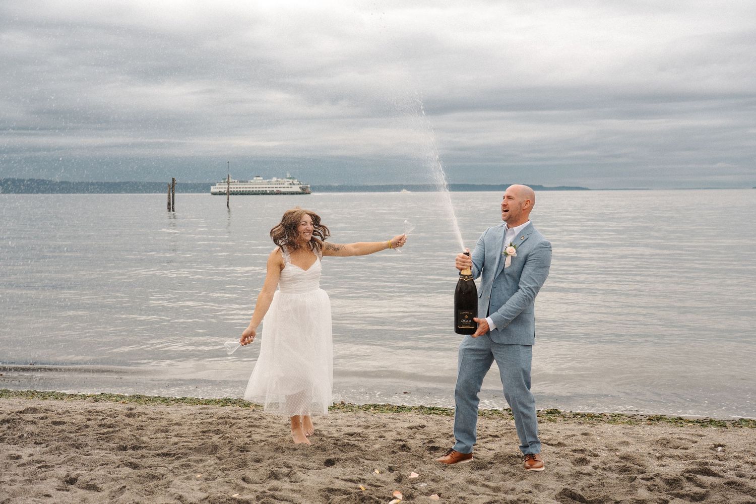 A bride and groom celebrate with champagne while walking along a misty beach on their wedding day.