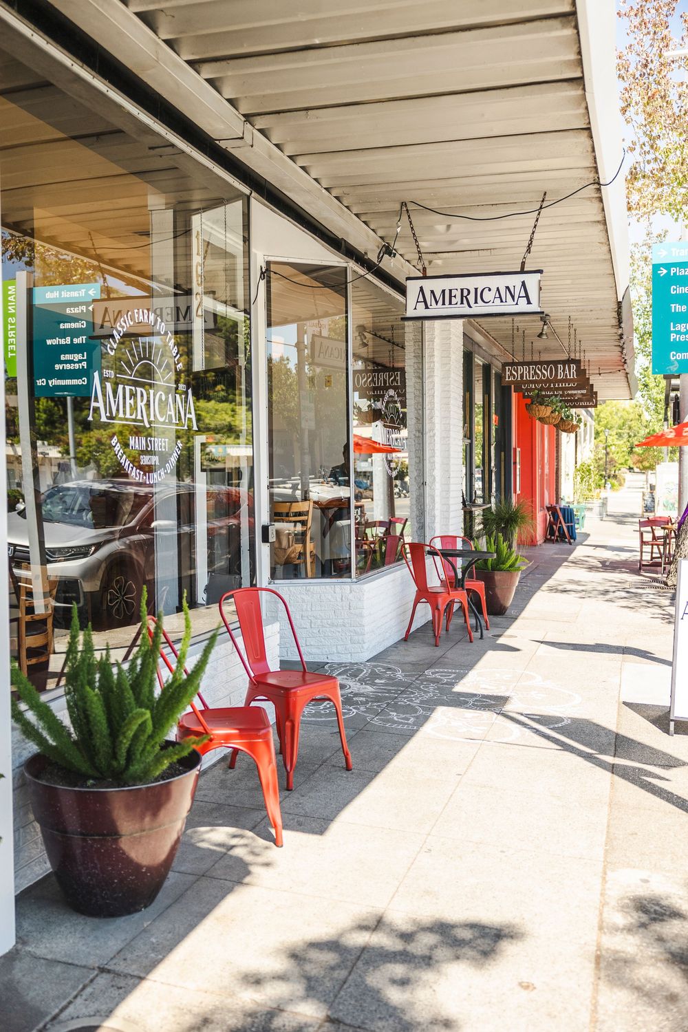 Storefront of Americana restaurant with red metal chairs and potted plants on a sunny sidewalk in downtown area.