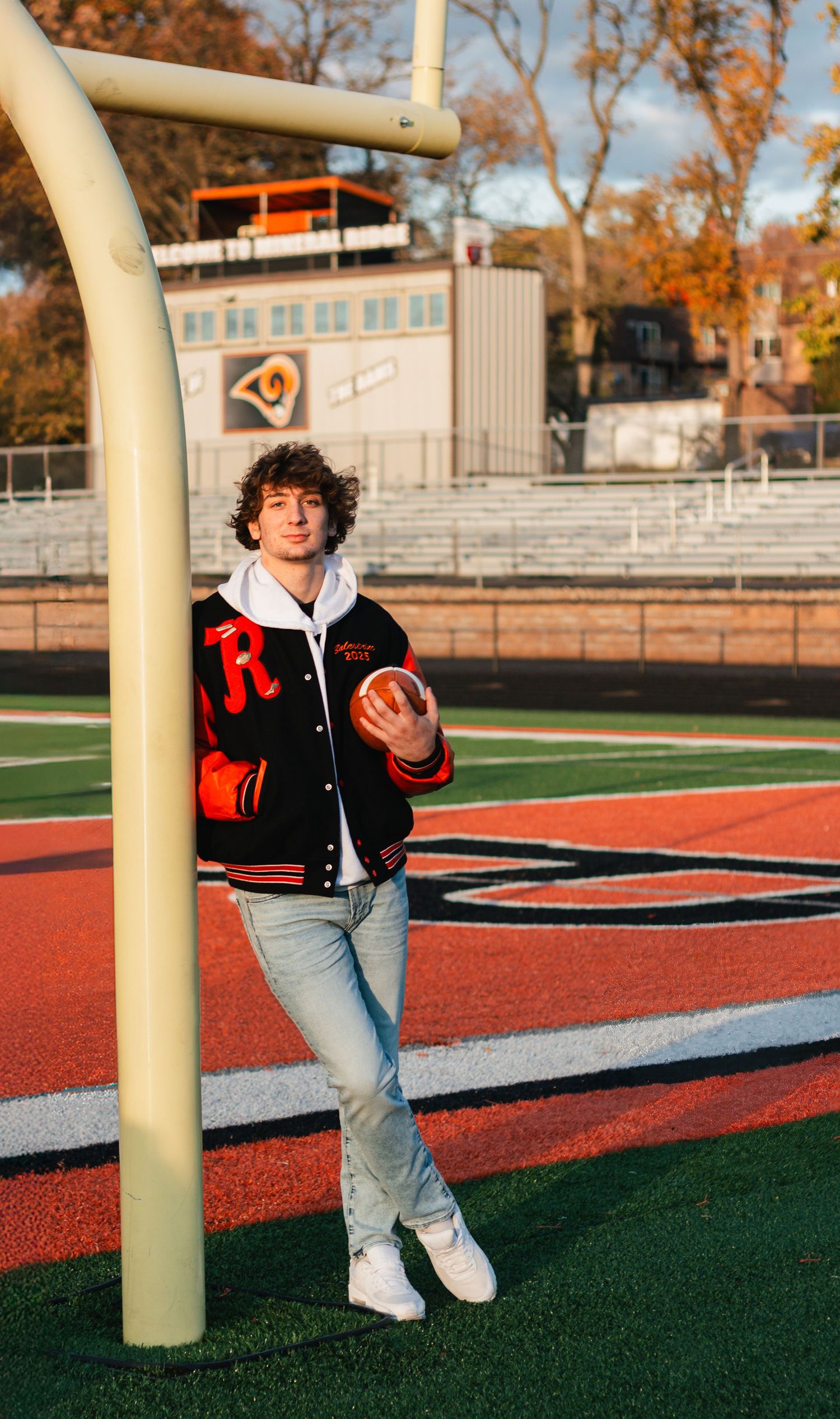 Varsity letterman stands on football field holding ball near goal post during autumn afternoon.