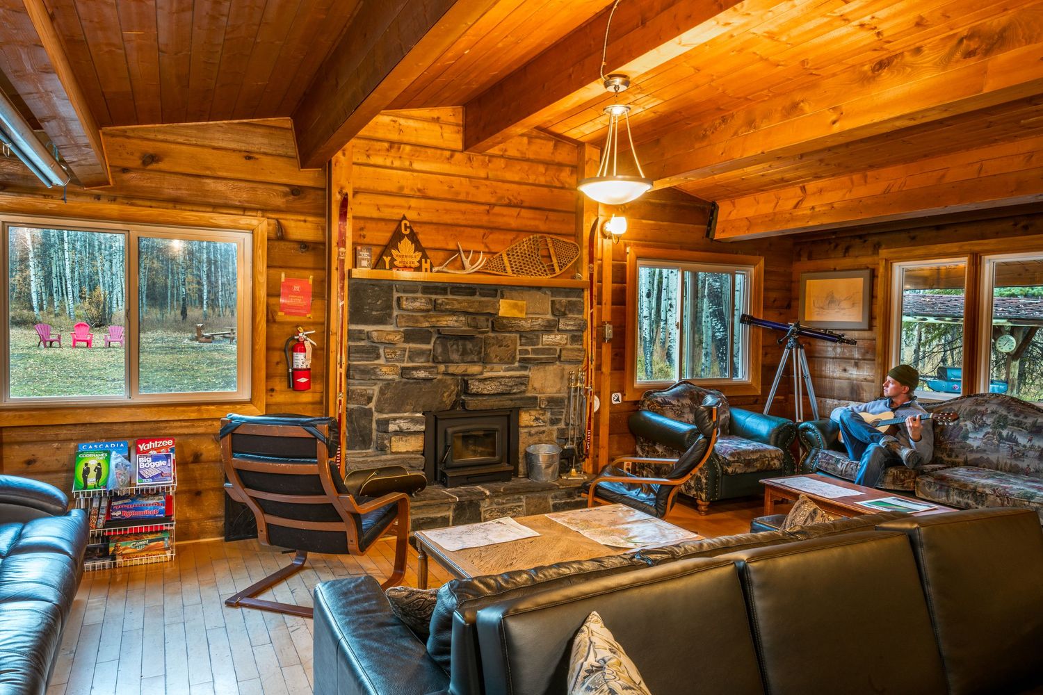 Cozy log cabin interior with stone fireplace, wooden beams, leather furniture and large windows overlooking forest views.