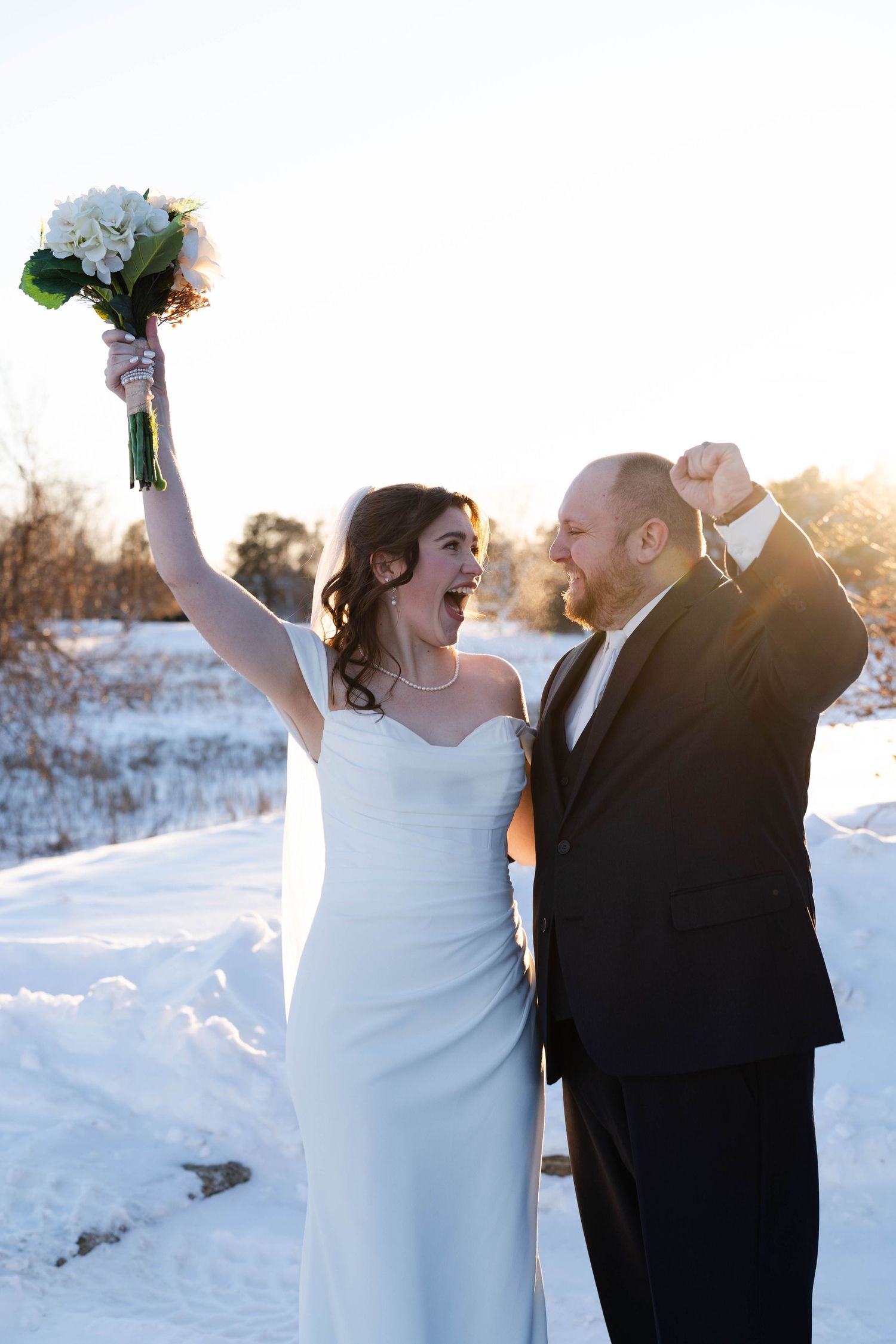 Joyful newlyweds celebrate outdoors in a snowy winter landscape at sunset with raised arms and bouquet.