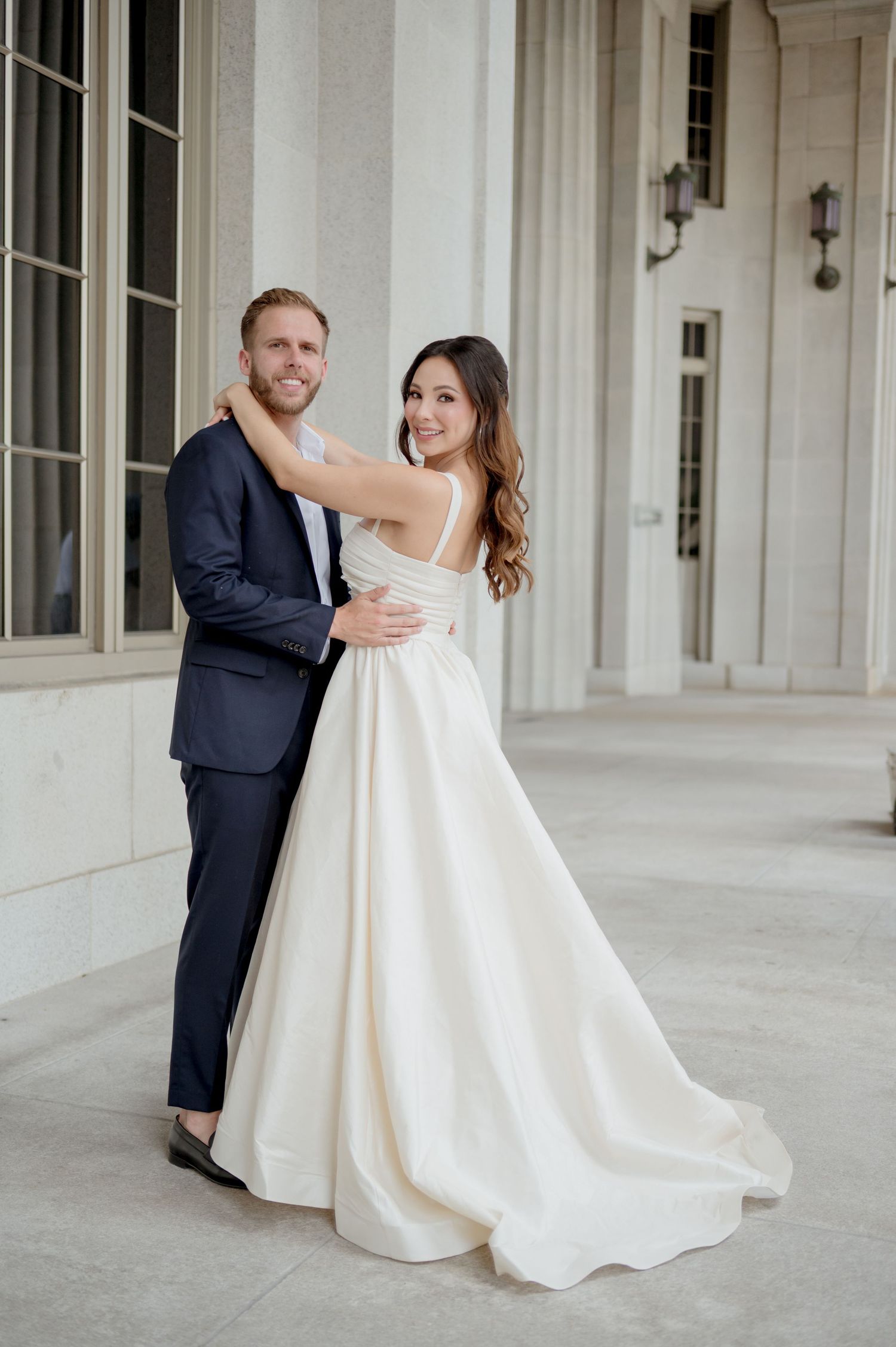 A bride in a flowing white gown dips gracefully while dancing with her partner in a navy suit on an elegant porch.