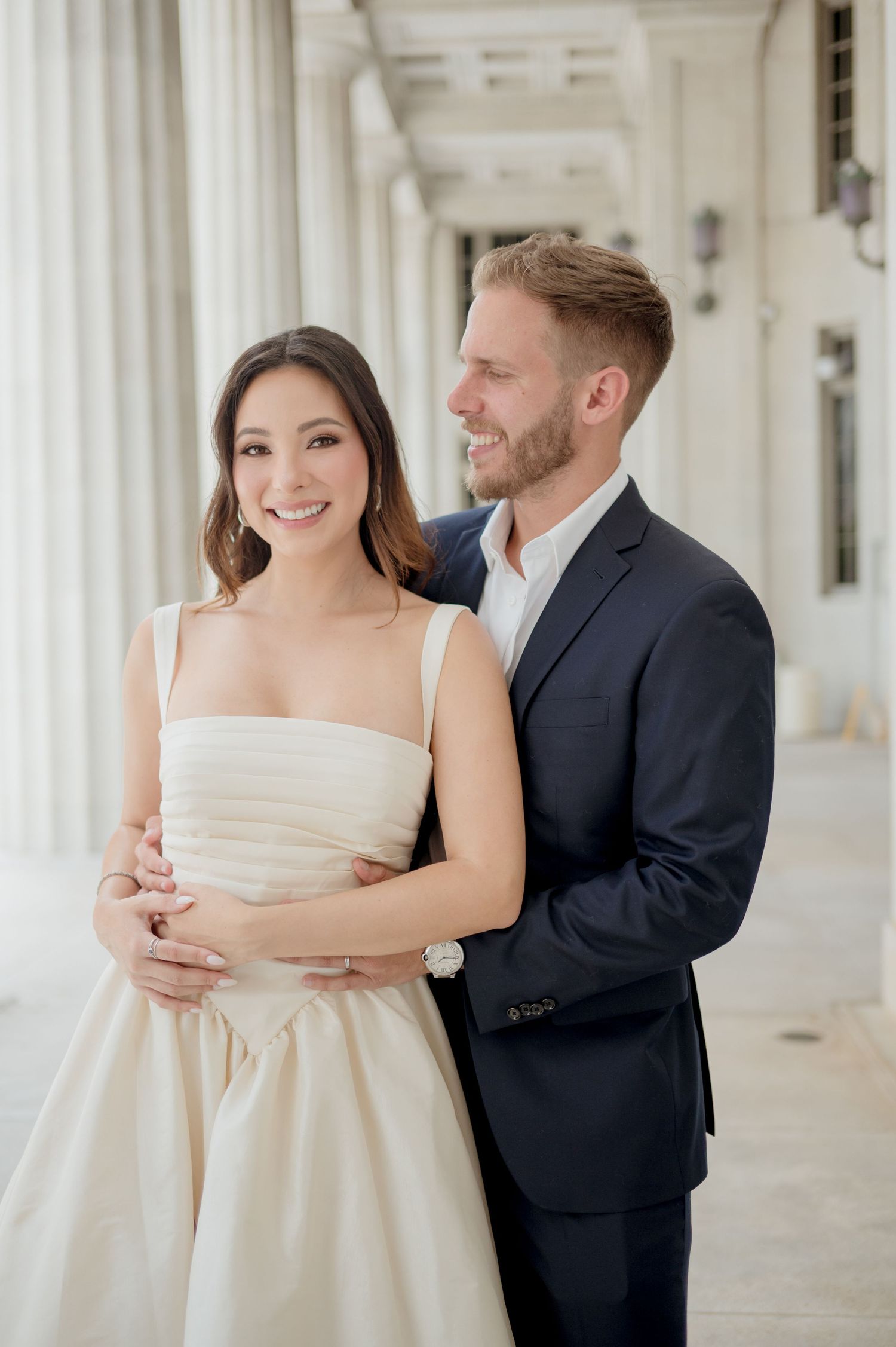 A joyful couple in wedding attire embraces and shares romantic moments along a pillared white corridor.