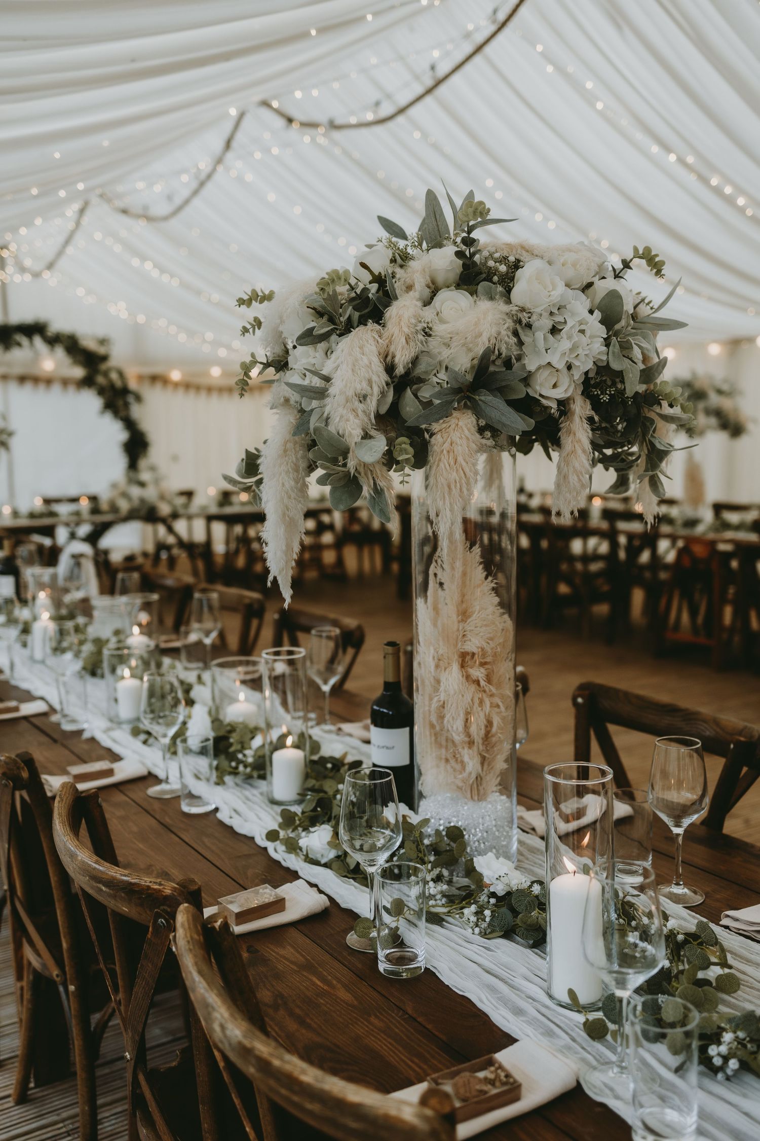 Elegant wedding reception table setting with white floral centerpiece and rustic decor under draped tent with twinkle lights.