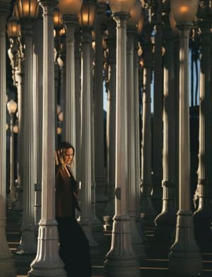 Dramatic photo of someone in black clothing standing among tall white architectural columns with warm lighting.
