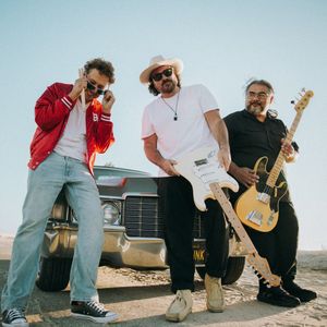 Musicians in casual attire perform outdoors with guitars and instruments against a desert landscape backdrop.