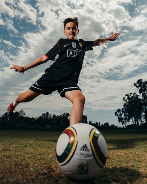Young soccer player in black Manchester United jersey kicking an Adidas ball on a grass field under a cloudy sky.