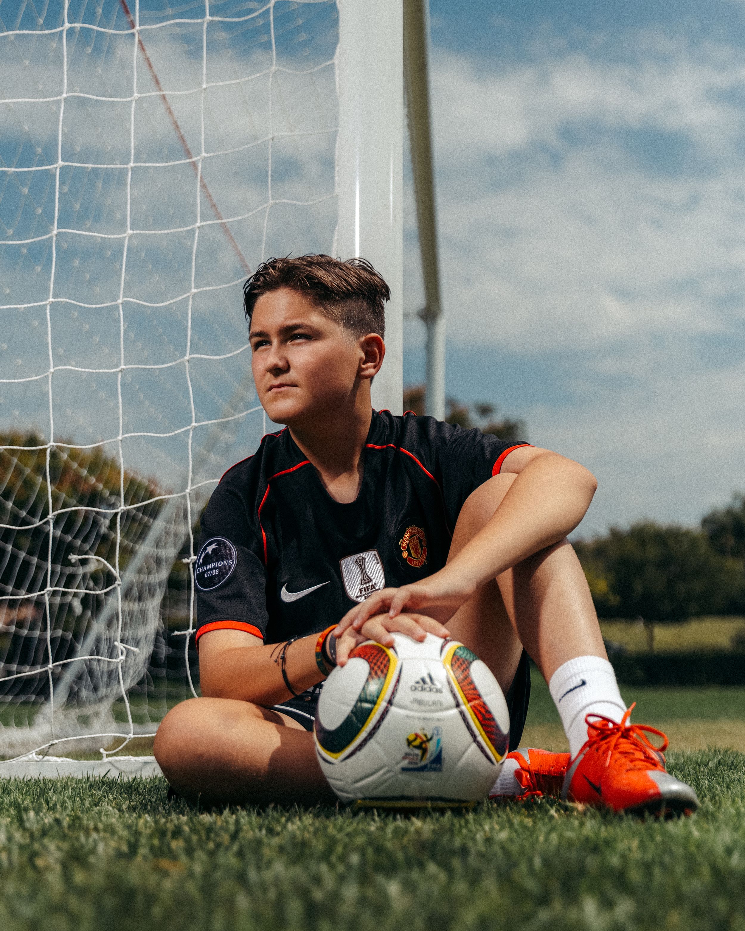 Young soccer player sitting by goal post on grass field, holding Adidas ball, wearing Nike jersey and orange cleats.