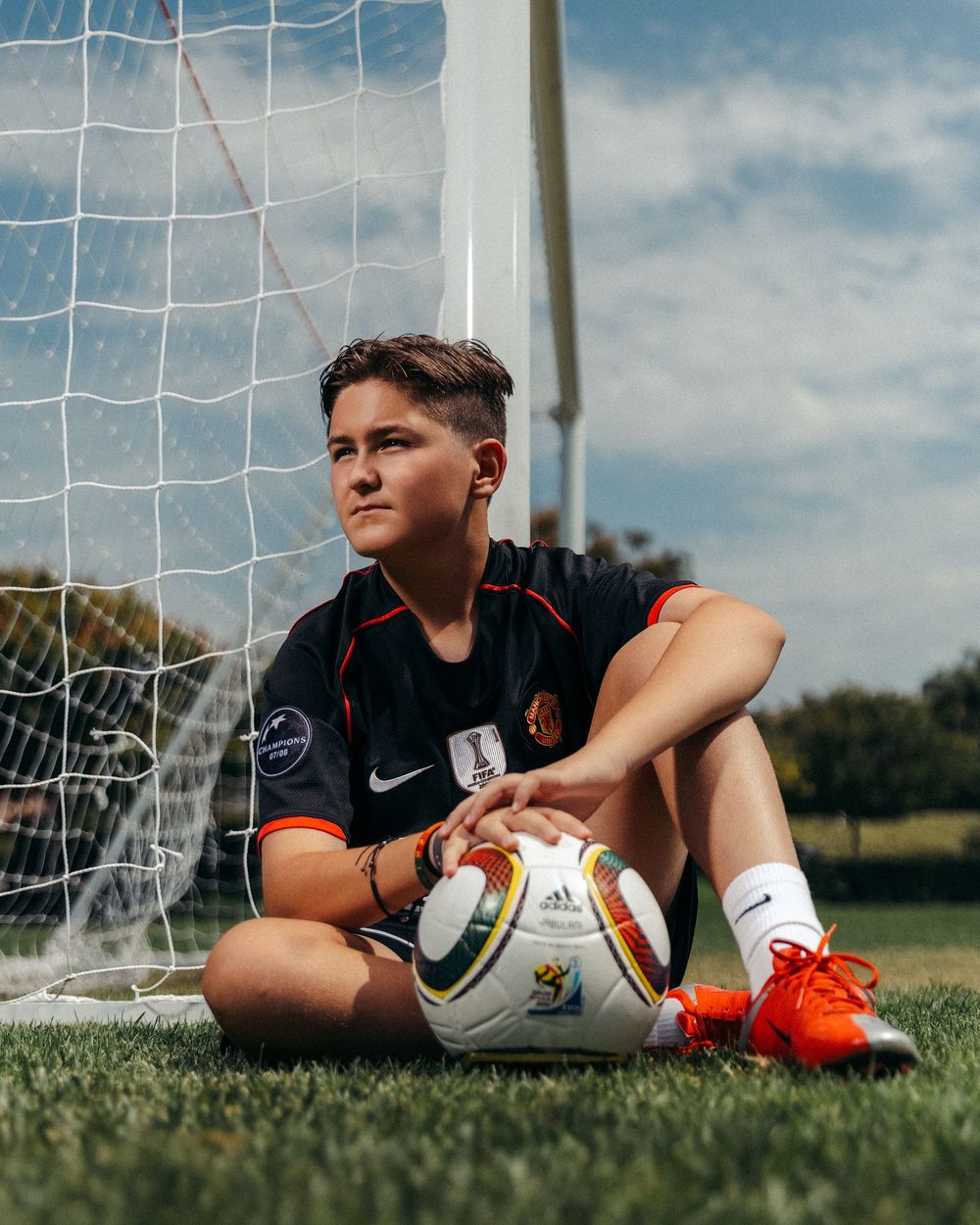 Young soccer player sitting by goal post on grass field, holding Adidas ball, wearing Nike jersey and orange cleats.