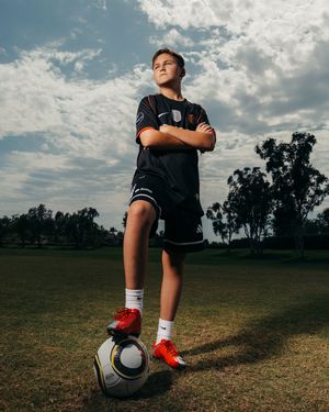 Young soccer player stands confidently with foot on ball, arms crossed, wearing black kit and red cleats on green field.