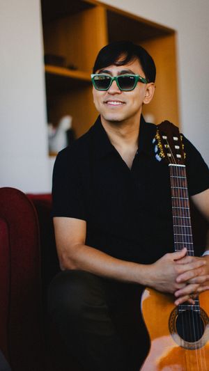 Musician in sunglasses and black polo shirt smiles while holding acoustic guitar against warm-lit background.
