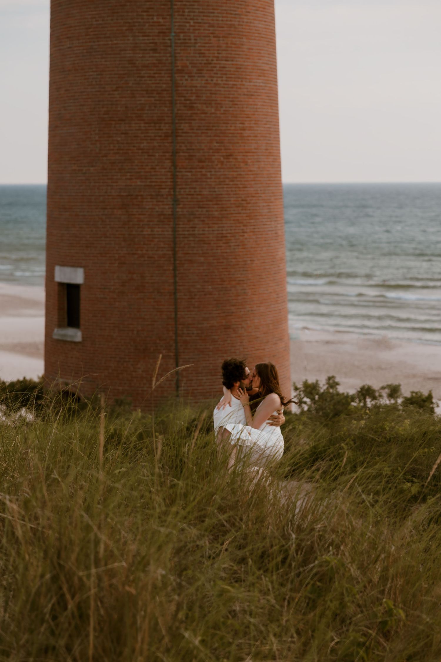 A romantic couple embraces near a tall brick lighthouse beside the ocean at sunset, surrounded by wild grasses. Rachel Martinez Photography