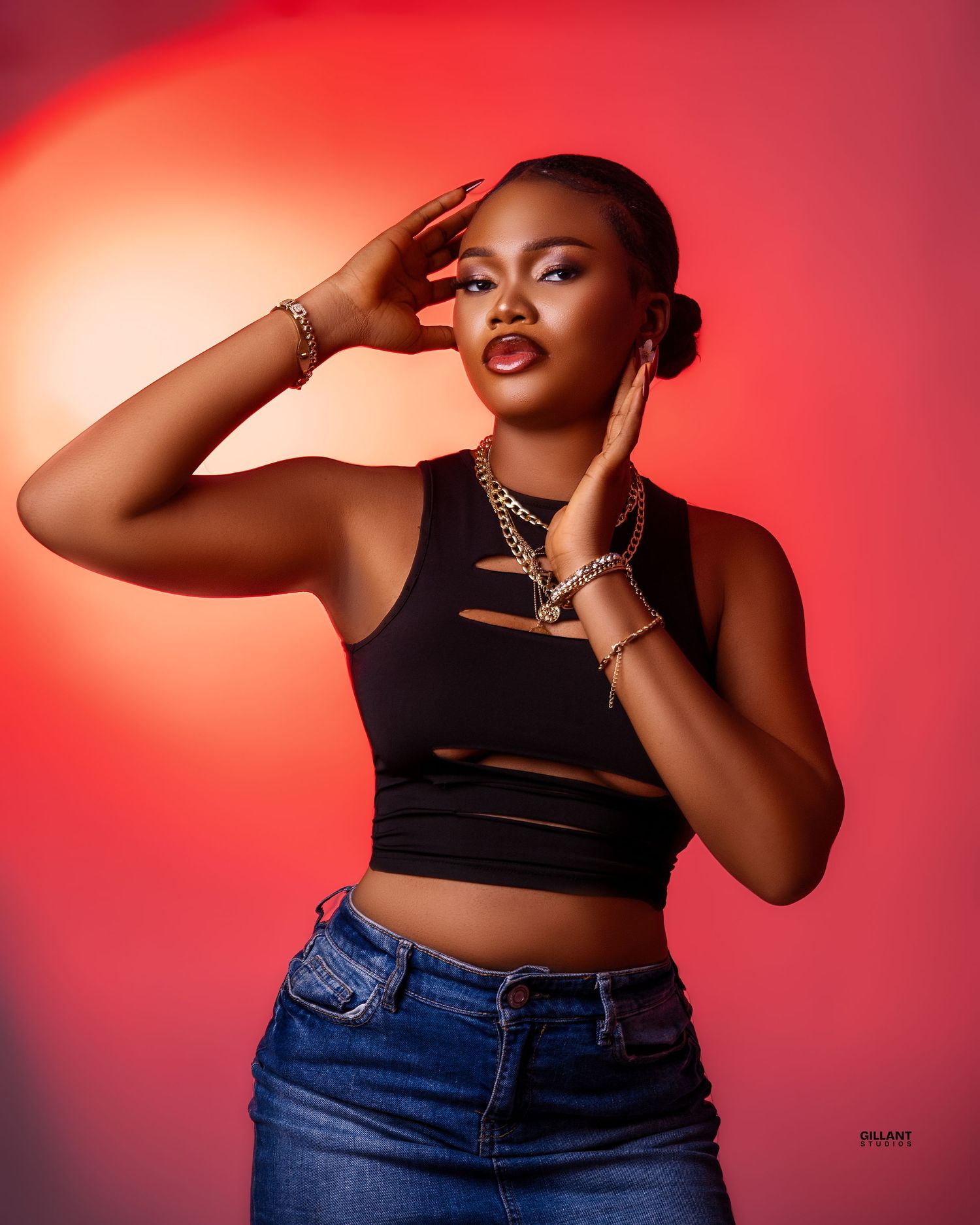 Dramatic portrait against red backdrop featuring black crop top and denim with jewelry accessories.
