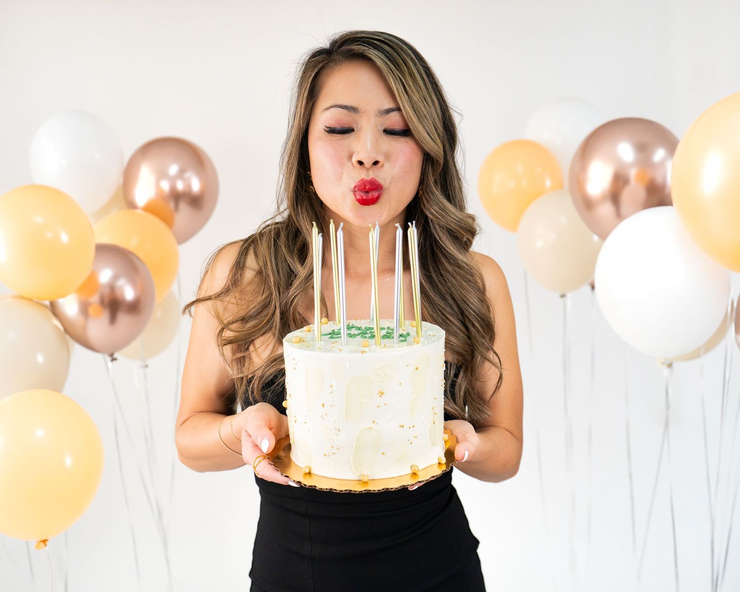 Woman in black dress holding a white cake with tall candles, framed by gold and white balloons.