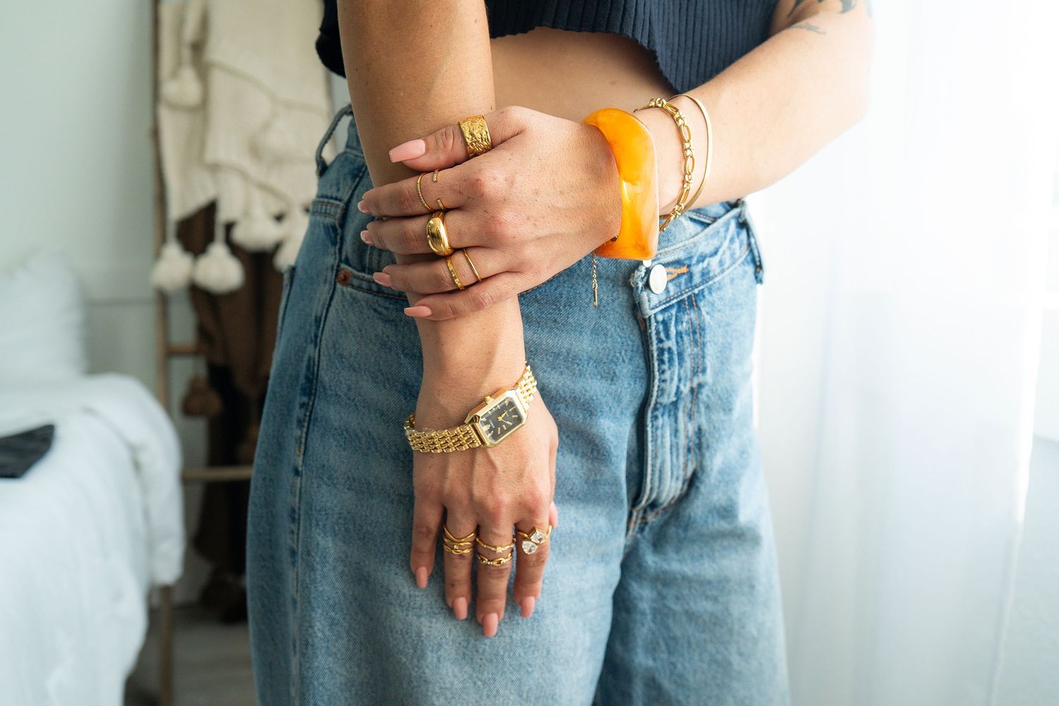 Close-up of person in jeans and crop top wearing layered gold rings, watch, and orange bangle.