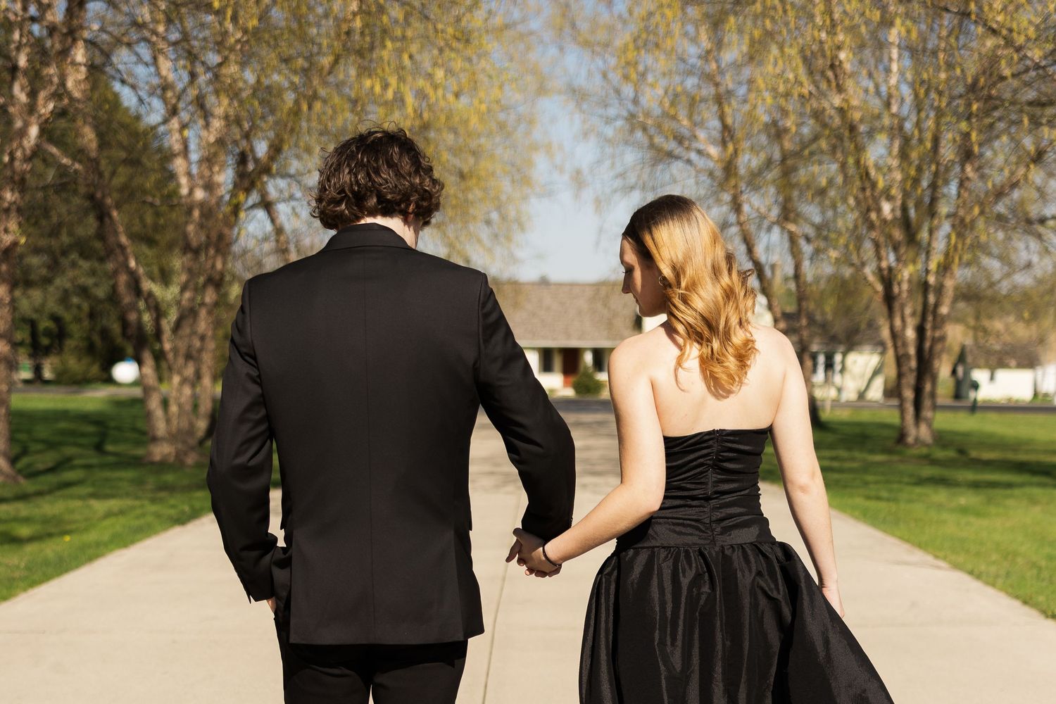 A couple in formal attire walks down a tree-lined path, the woman wearing a black dress and man in a dark suit.