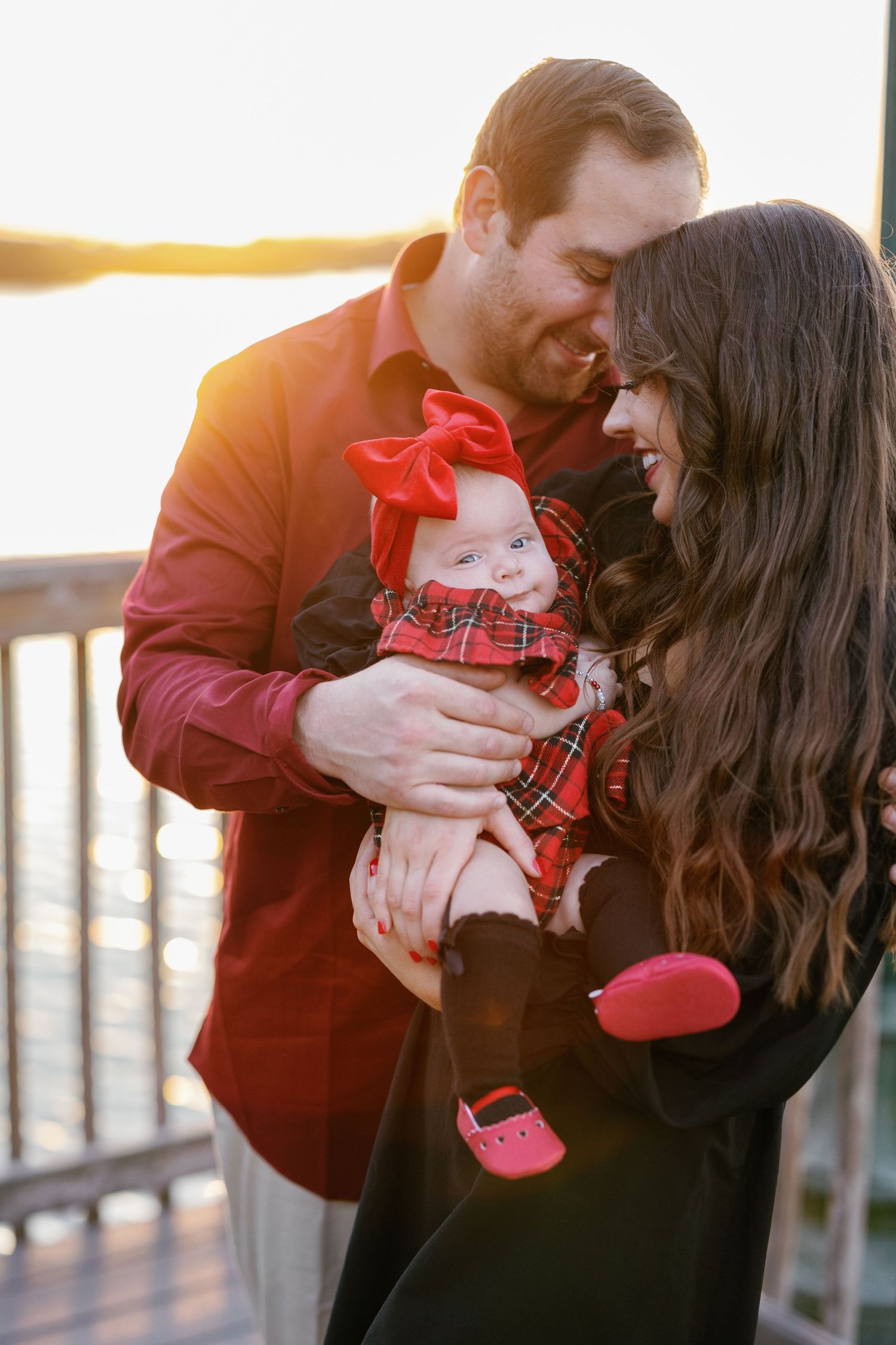 Happy family sharing tender moment outdoors at sunset with baby wearing red bow and plaid outfit on wooden deck.