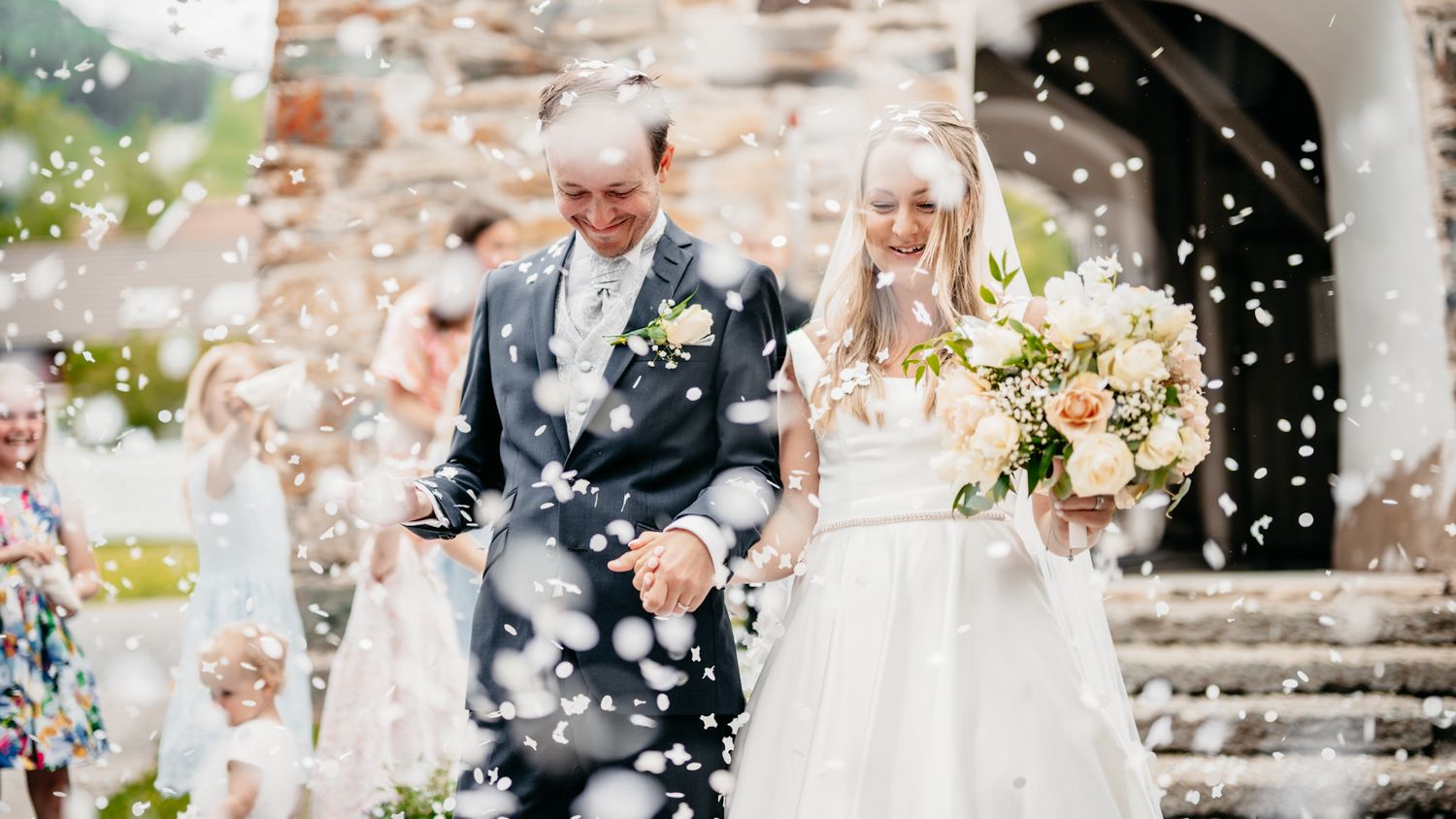 Joyful wedding celebration with confetti toss outside historic stone building during outdoor romantic ceremony.