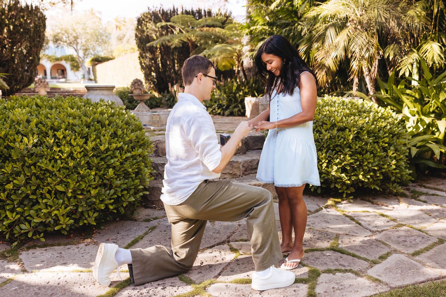 A romantic proposal scene outdoors at sunset with stone steps and lush gardens in the background.