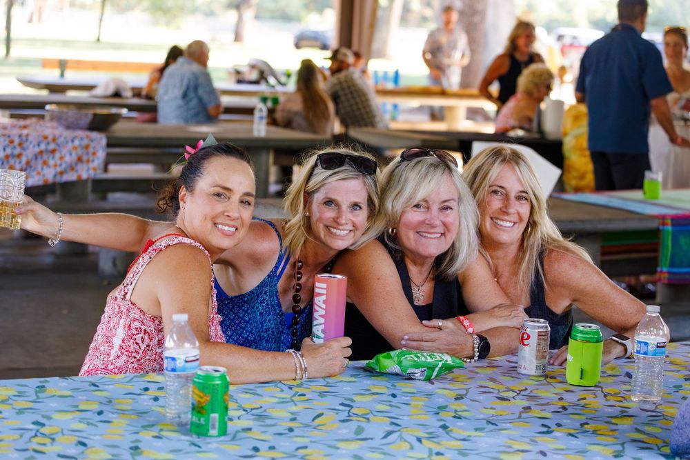 Four women smiling and posing together at a picnic table outdoors at a sunny summer gathering.