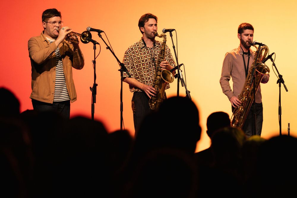 Three musicians performing with brass instruments against warm-colored backdrop.