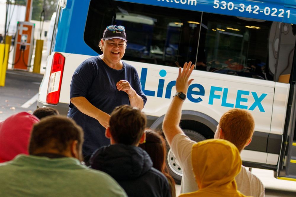 A transit educator engages a group of students in front of a B-Line FLEX bus during an educational field trip.