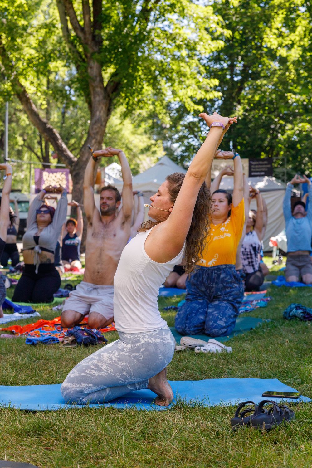 Group outdoor yoga class on grass mats in sunny park, woman in white leading pose with arms raised.
