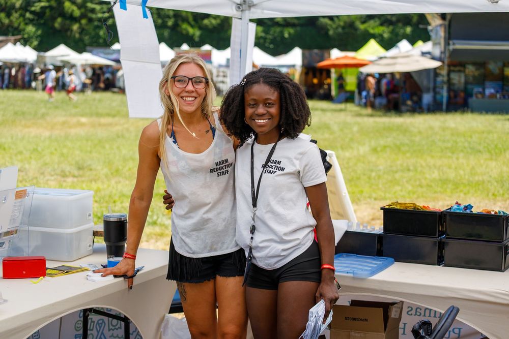 Two smiling women in matching white shirts stand together at an outdoor festival booth on a sunny day.