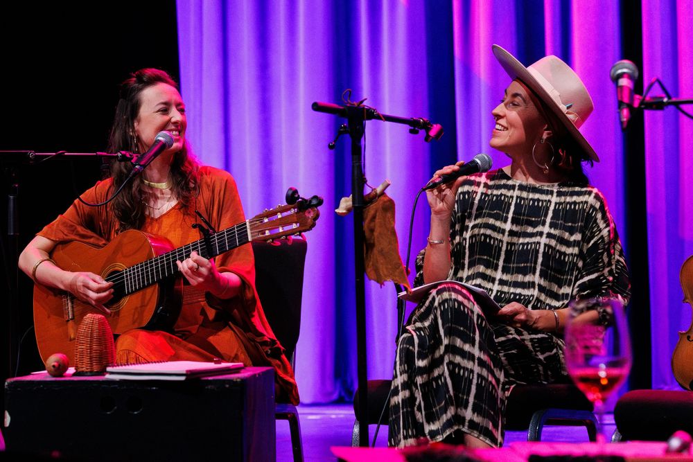 Two women performing live music on stage, one playing acoustic guitar in red, the other singing in a plaid dress.