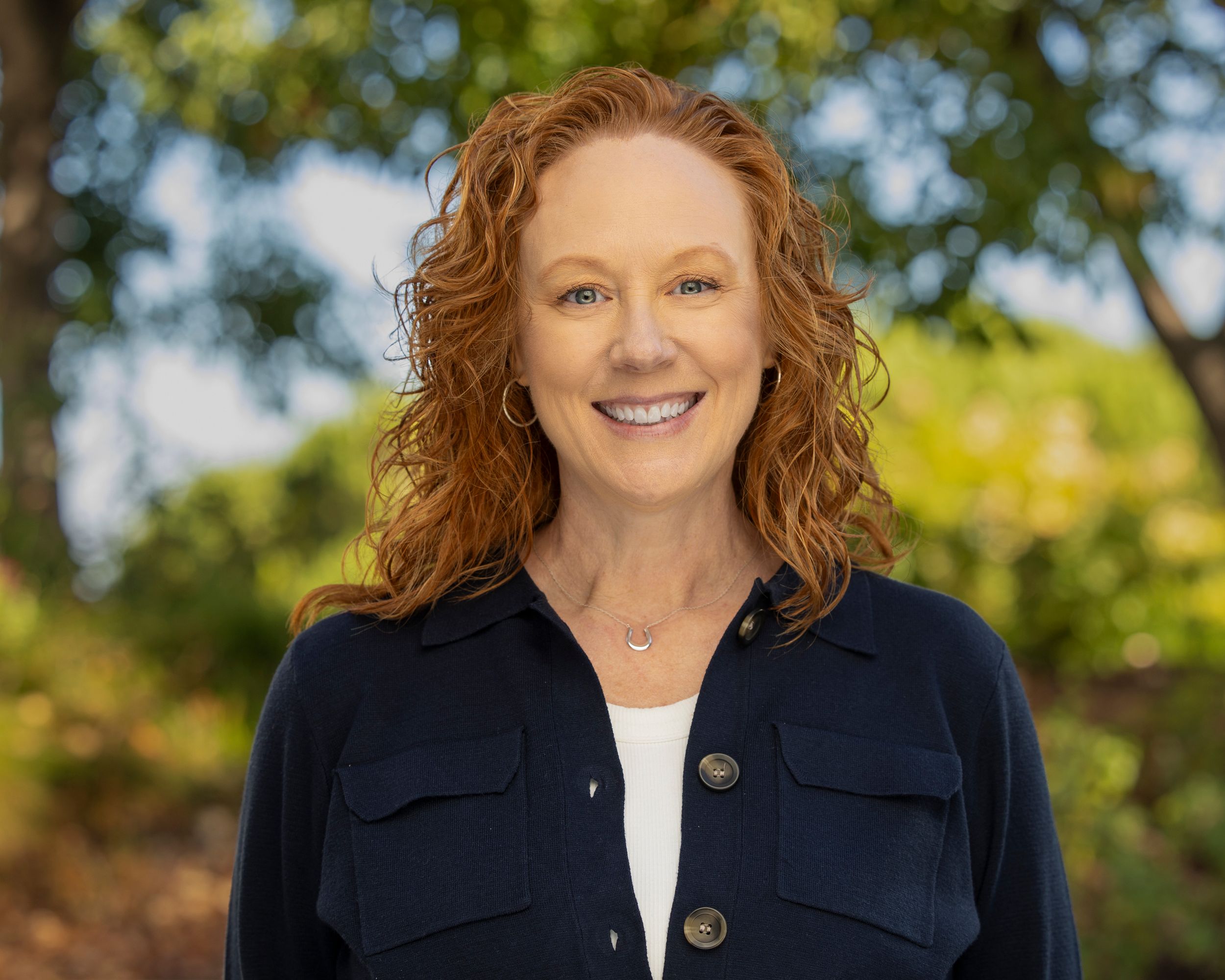 Professional outdoor portrait of someone wearing a navy button-up shirt and white top against a green natural background.