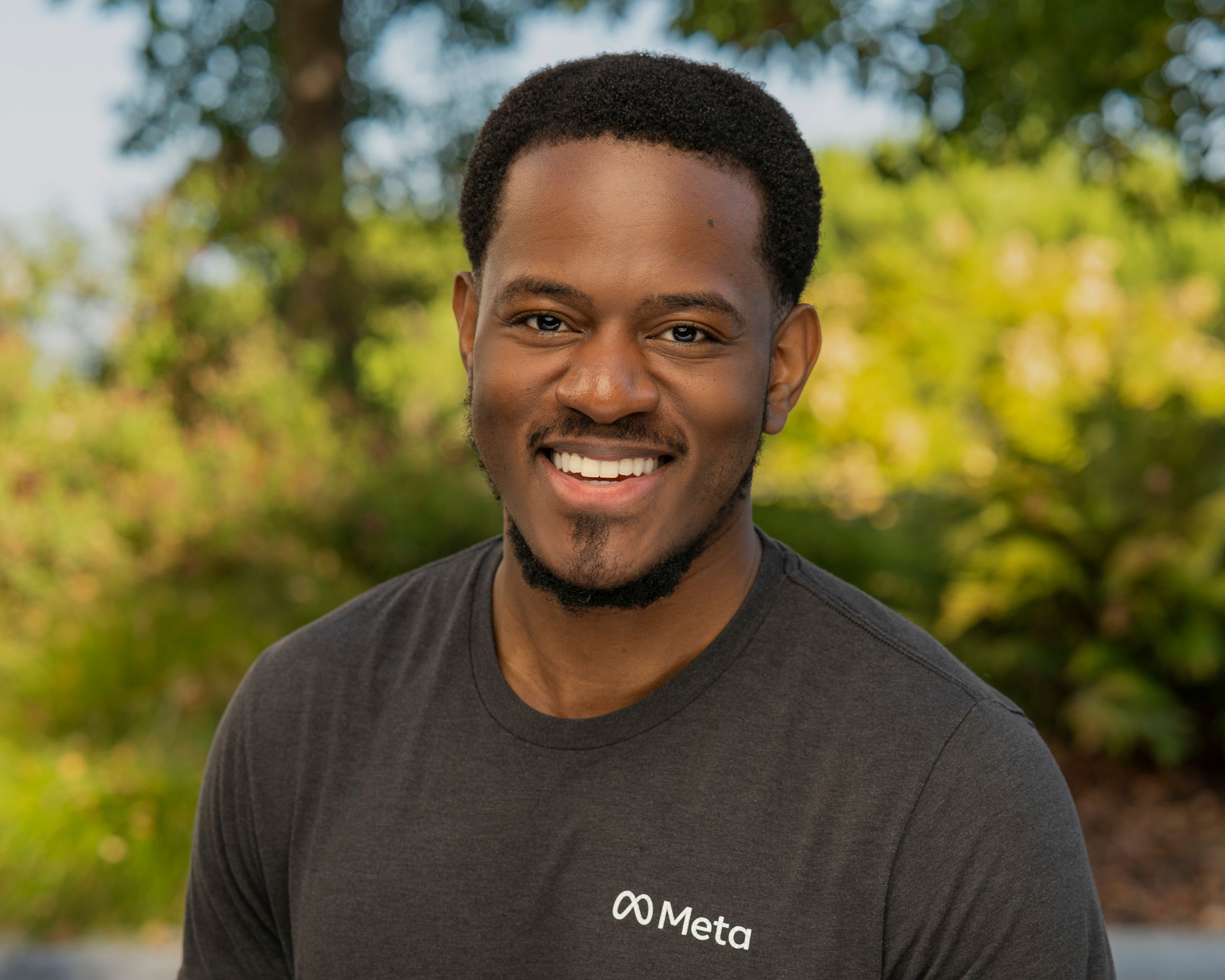 A person in a dark grey Meta t-shirt smiles outdoors on a sunny day with green foliage in the background.