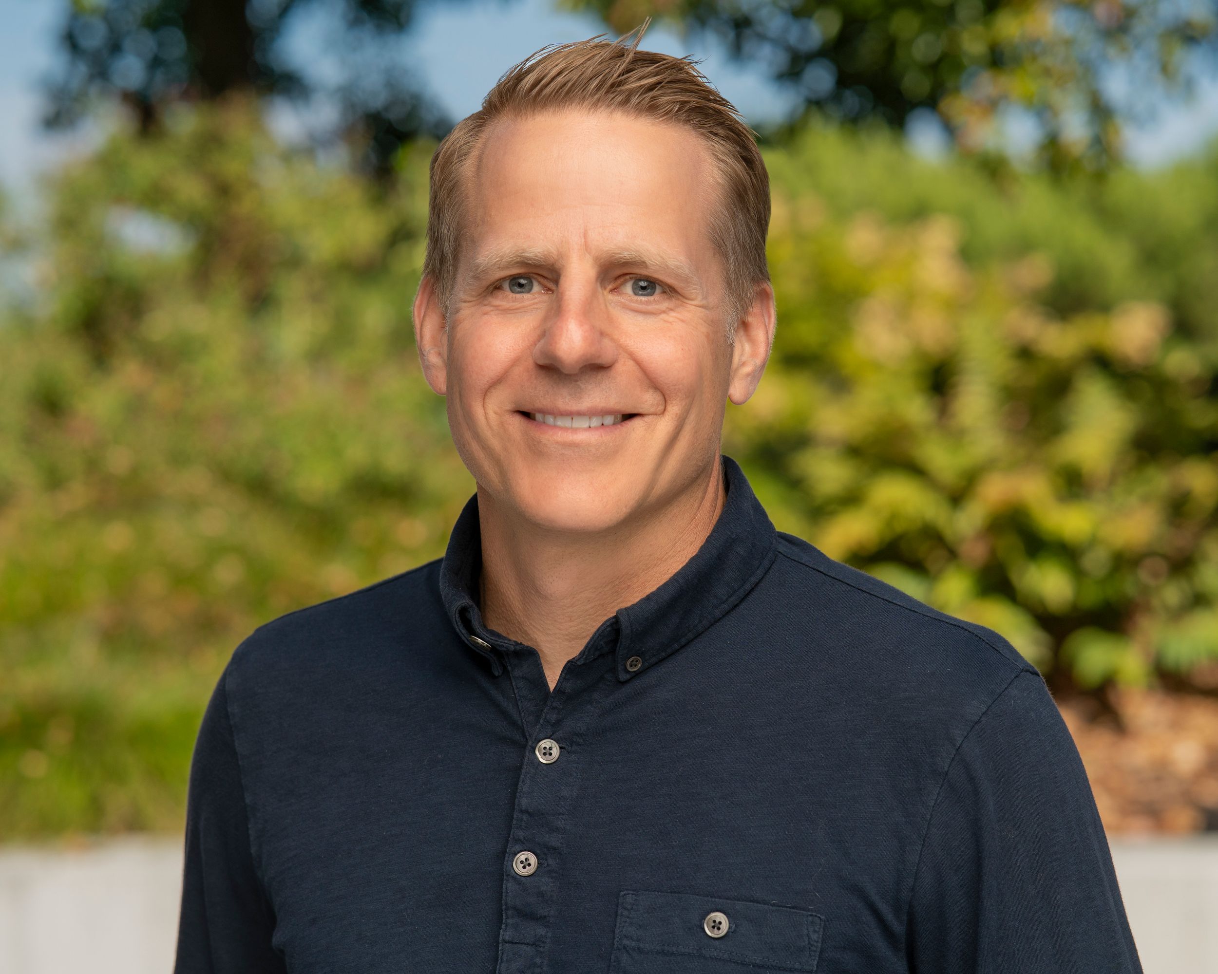 Professional headshot portrait of someone wearing a dark button-up shirt outdoors with greenery in the background.