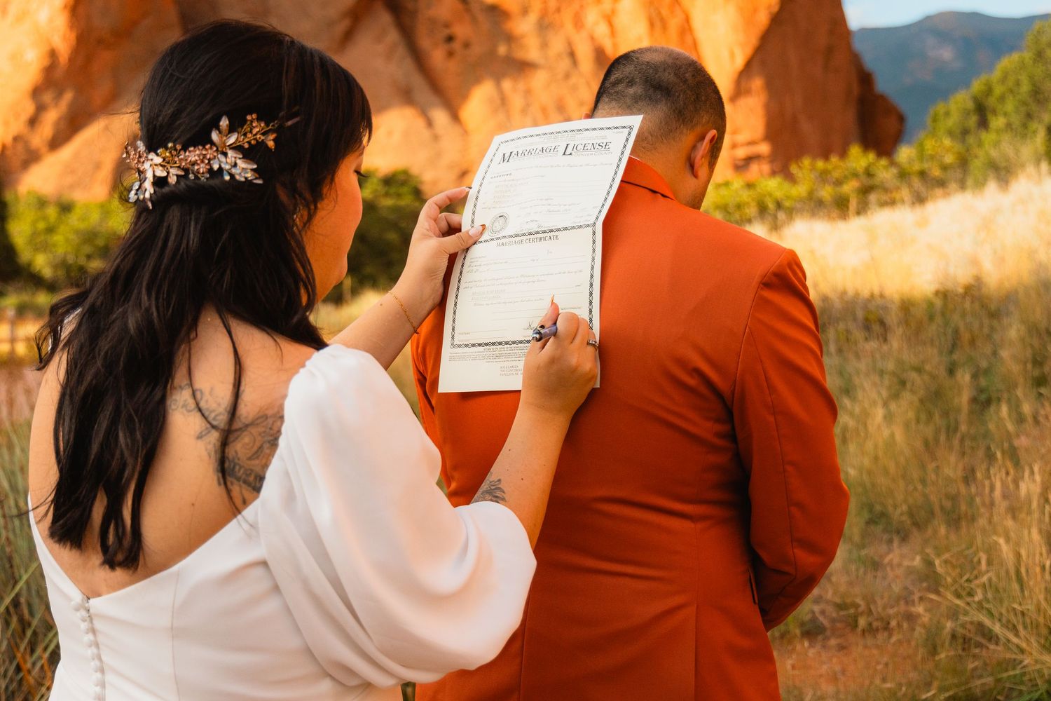 A couple in wedding attire signs marriage documents against a backdrop of rustic red rock formations at sunset.
