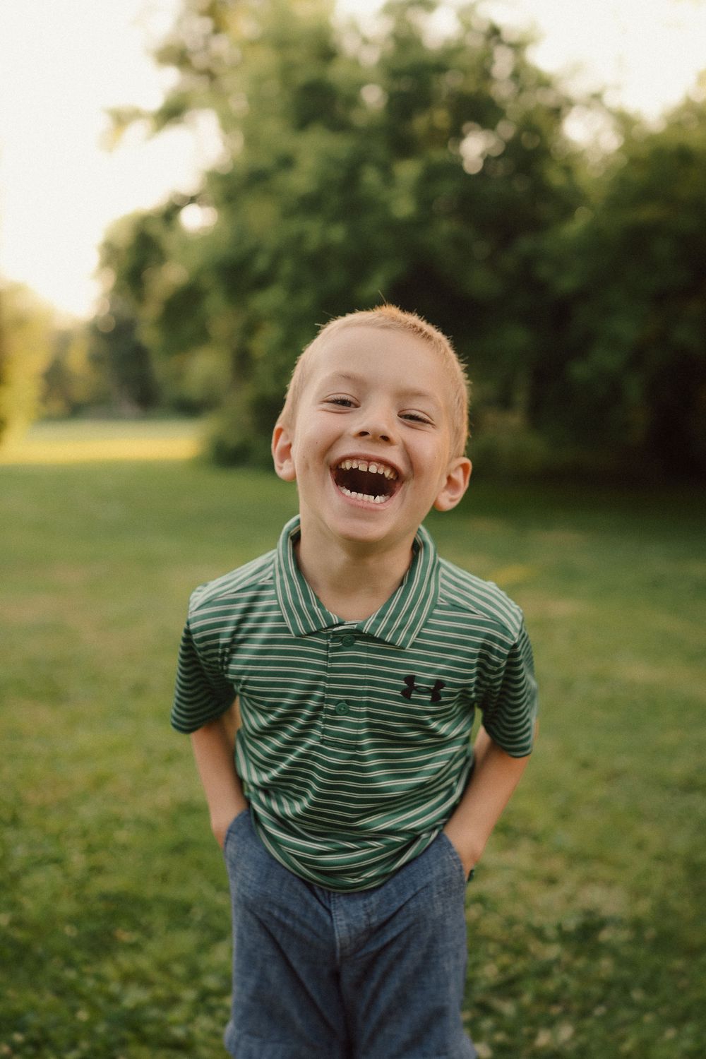 A young child in a striped green shirt and jeans plays joyfully in a grassy outdoor setting during golden hour.