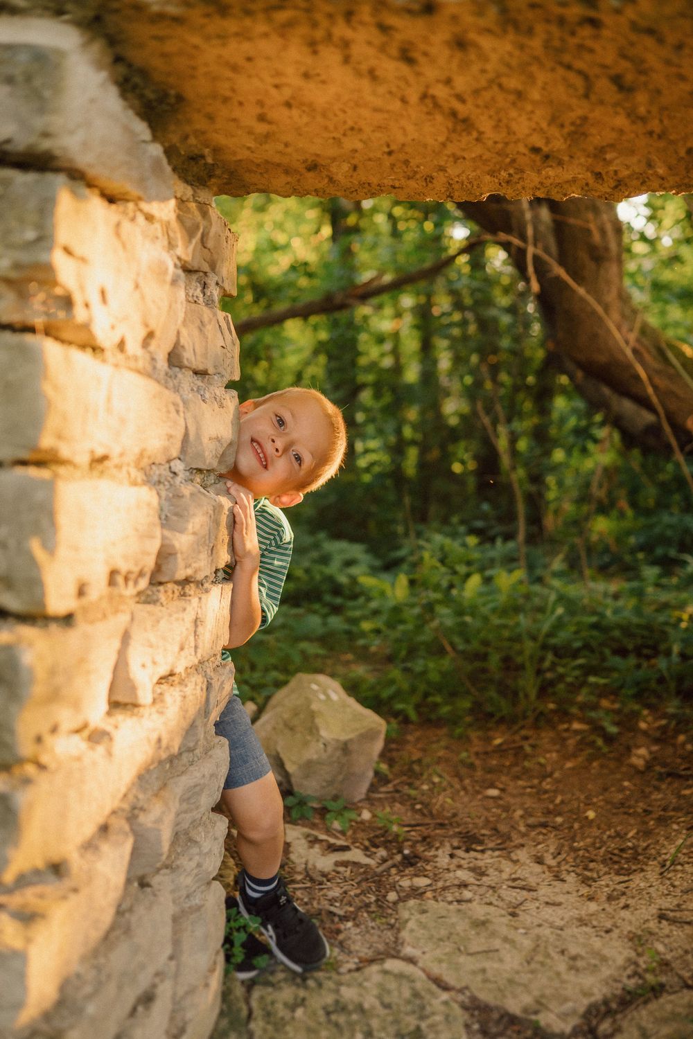A toddler in a striped shirt and shorts exploring a stone wall formation in a wooded area.