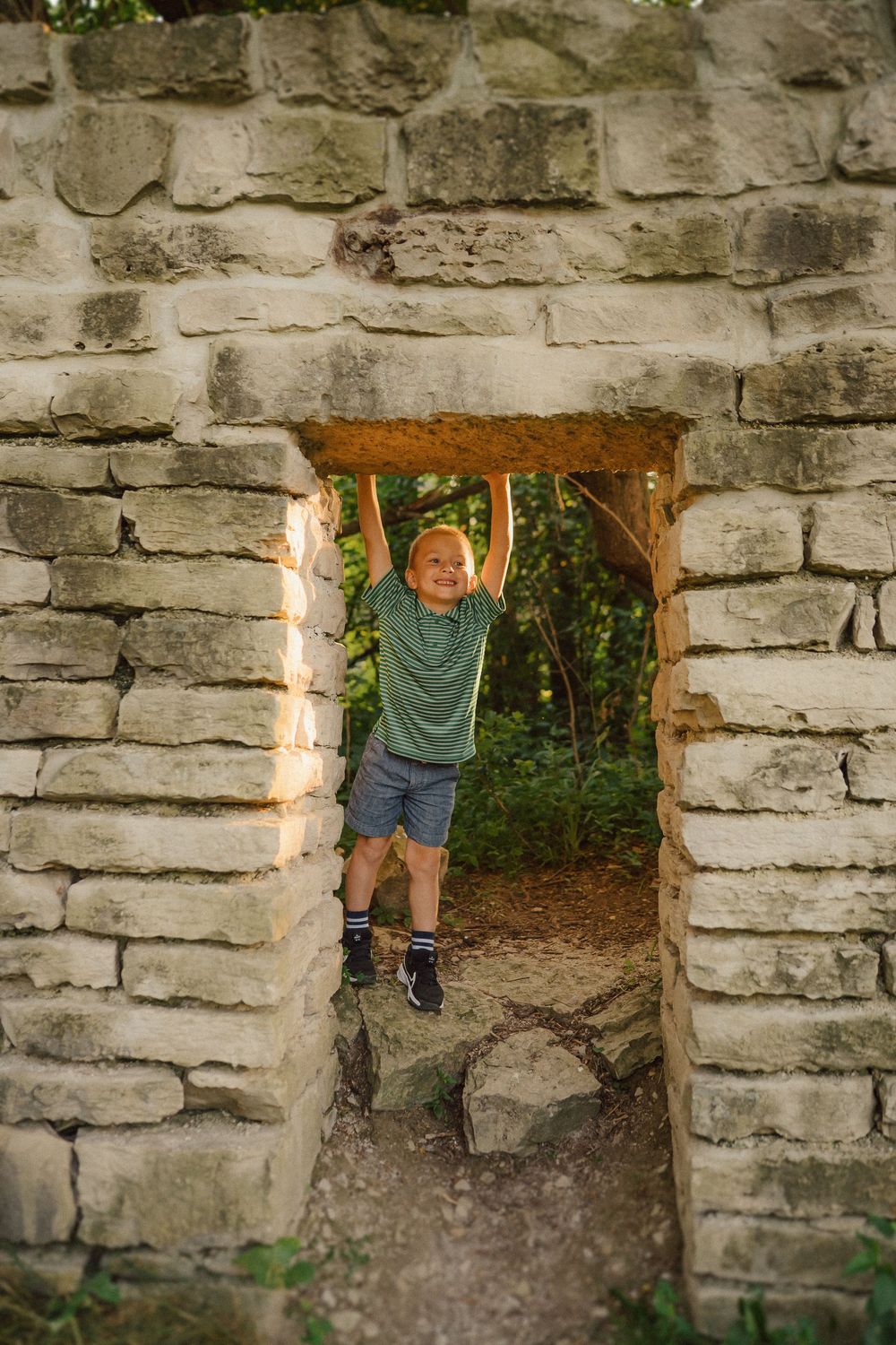 Child peeks playfully through openings in rustic stone wall during outdoor photoshoot.