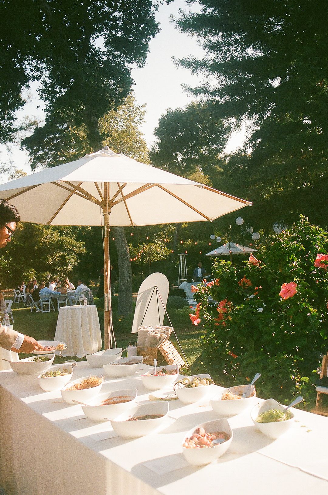 Outdoor buffet setup with white furniture and umbrellas under trees with sunlight filtering through the branches.