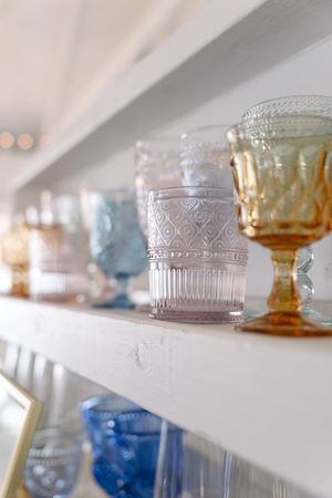 Vintage glass jars and decorative vessels with etched patterns displayed in a row on white shelves in soft lighting.