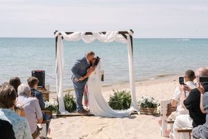 A romantic beach wedding ceremony takes place under a white draped arch as guests capture the moment with phones.