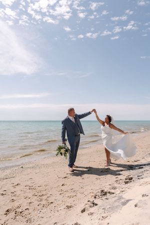 A couple shares a joyful moment on a sunny beach as a dress flows in the ocean breeze.