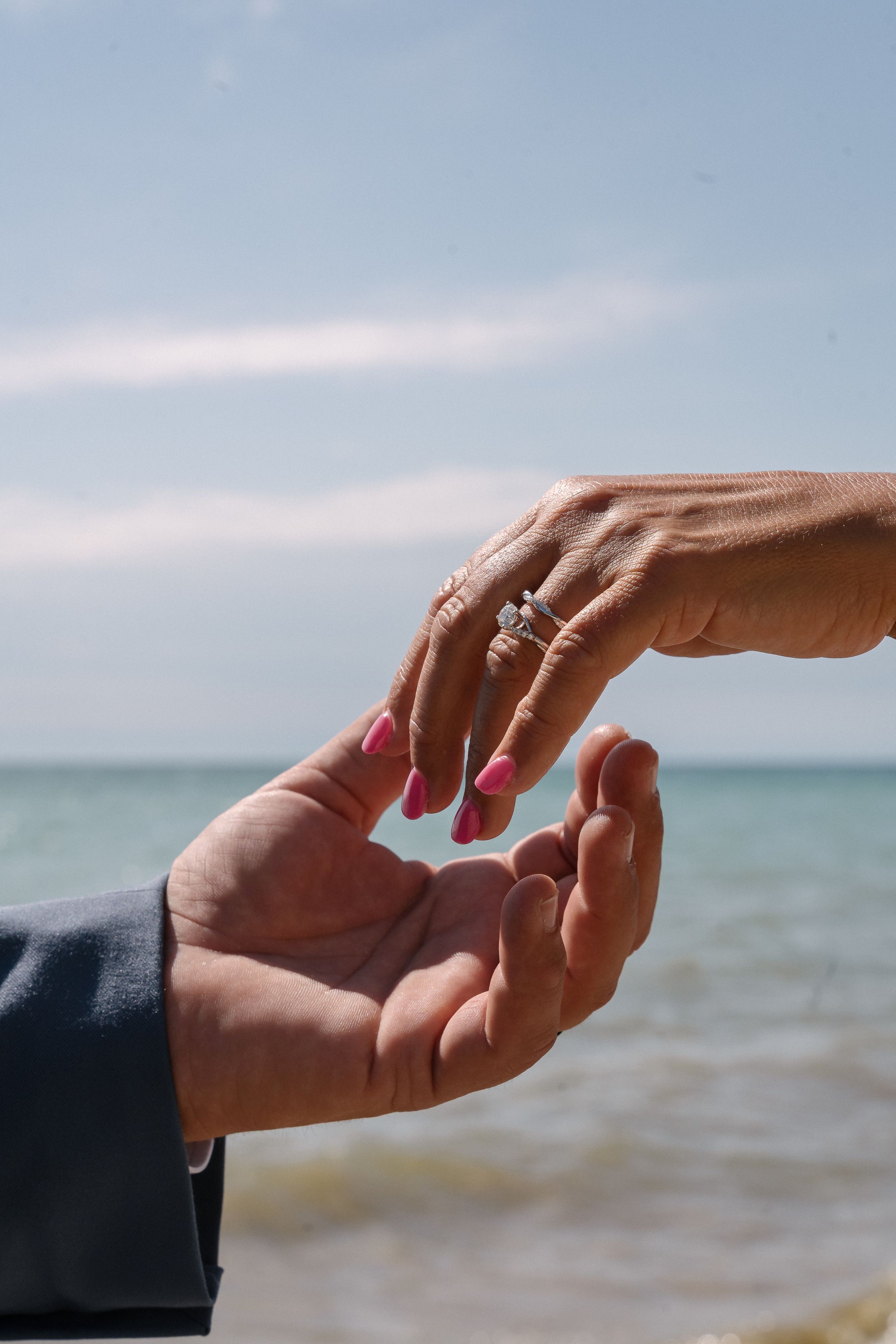 Two hands reaching towards each other with an engagement ring against a blurry ocean background.
