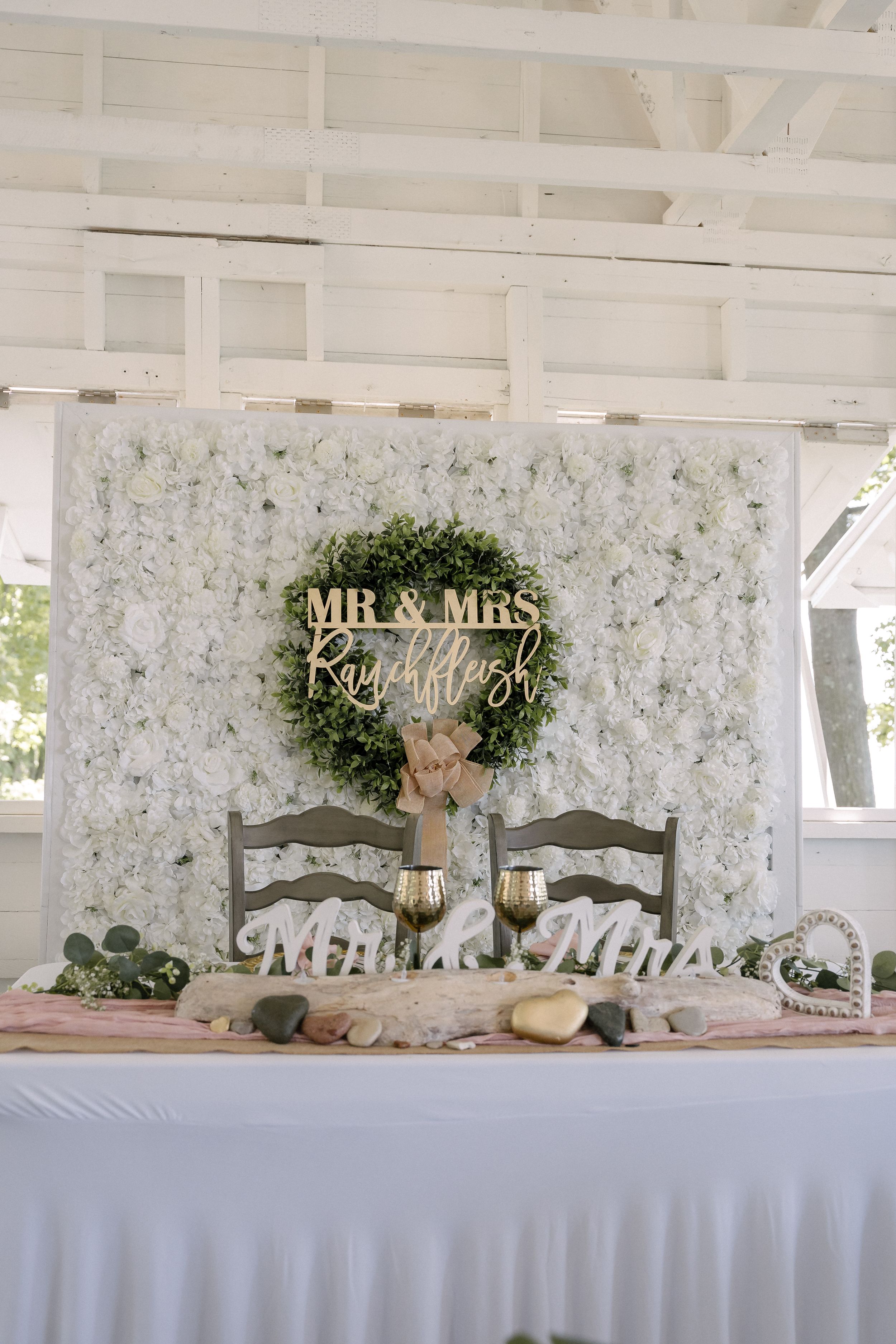 Elegant wedding sweetheart table with white floral backdrop, green wreath, Mr & Mrs sign and rustic wooden decor.