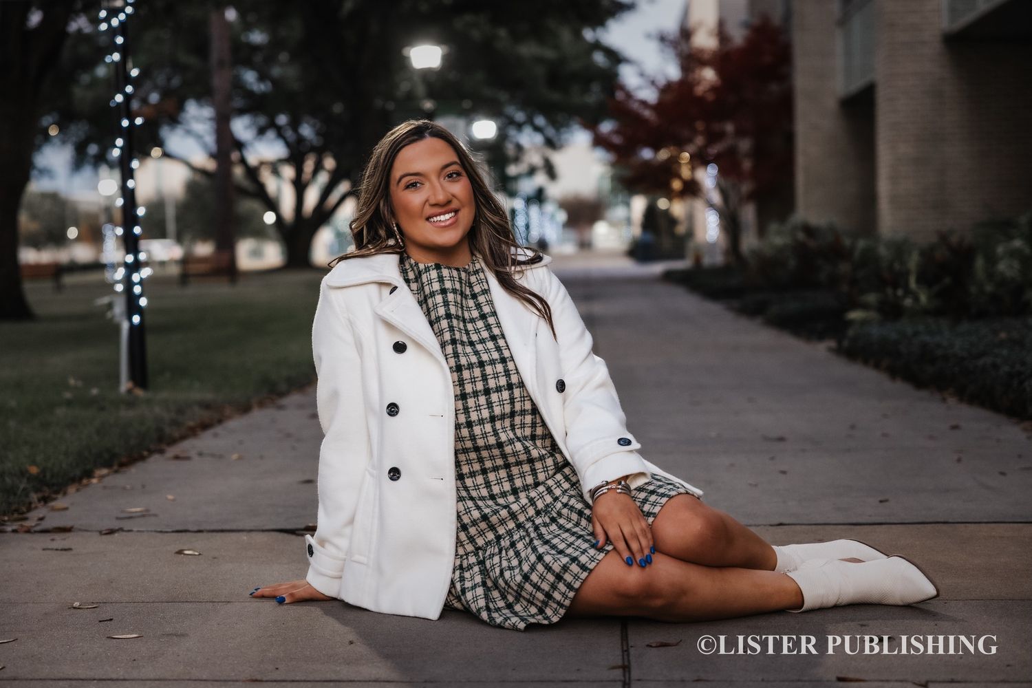 A person in a white peacoat and plaid dress sits on a sidewalk near twinkling holiday lights at dusk.