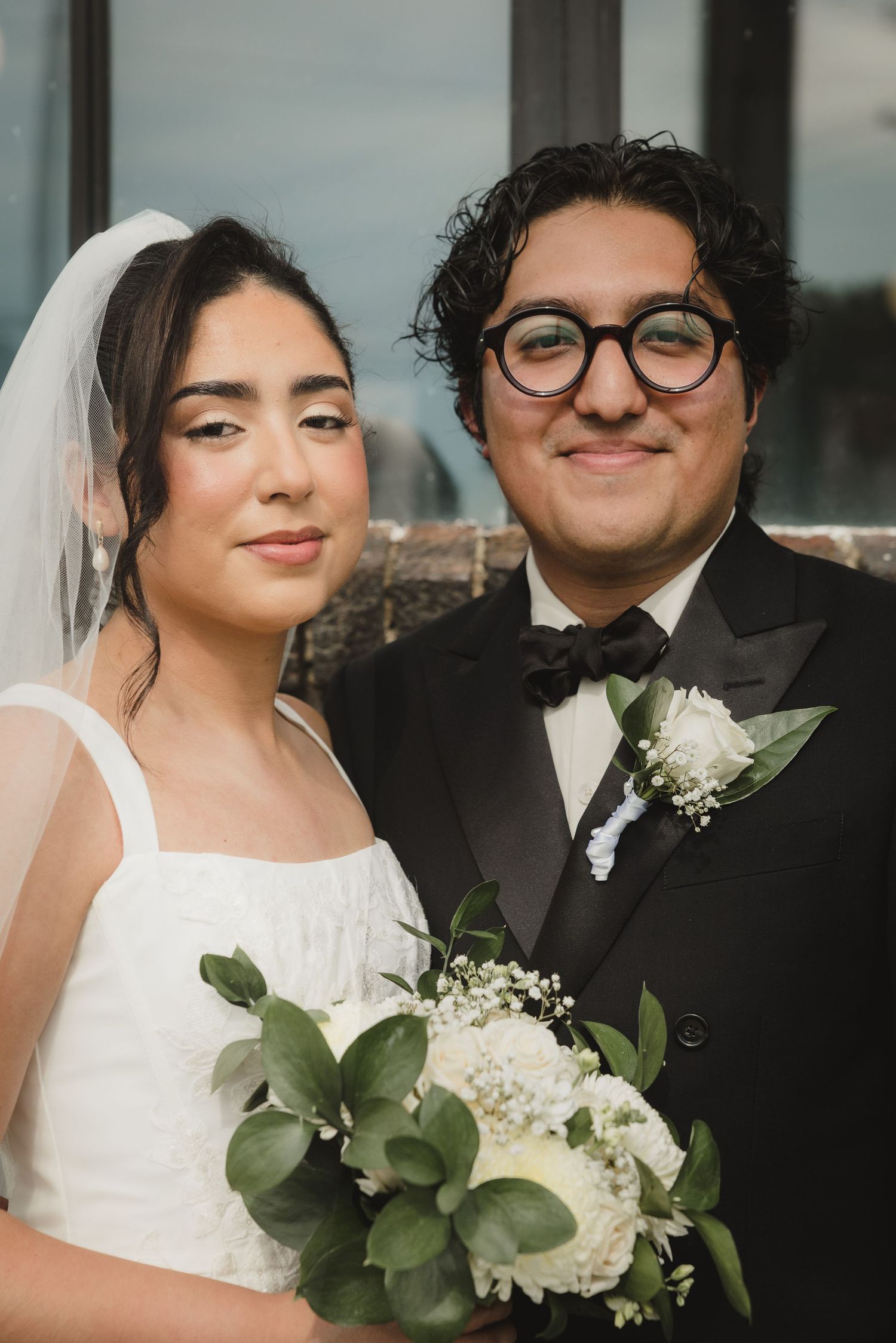 A bride and groom pose together on their wedding day, she holds white floral bouquet, he wears black tuxedo with boutonniere.
