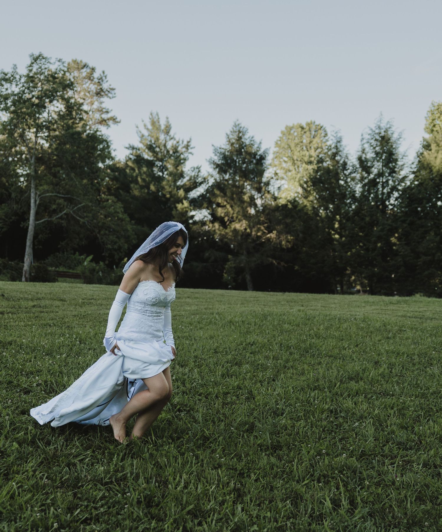 Bride in white wedding dress and veil walking barefoot on lush green grass with trees in background.