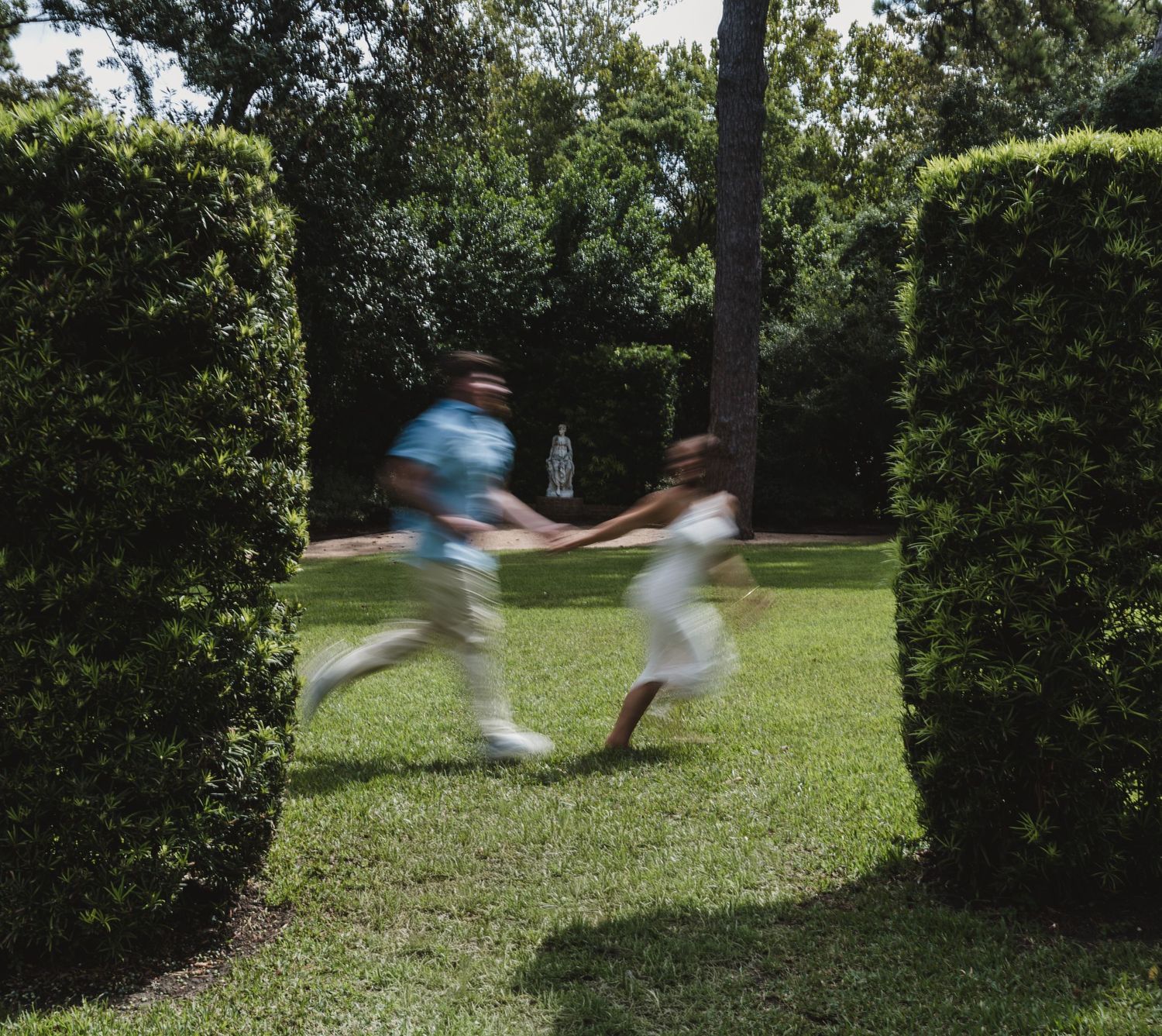 Two children running hand-in-hand through a garden pathway flanked by tall trimmed hedges on a sunny day.